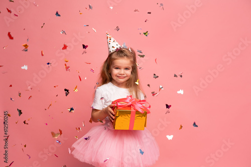 little girl in party hat holds gift box in front of a pink background with confetti