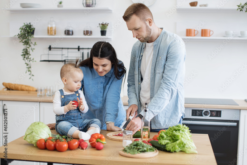Delighted family man feeding little daughter while smiling mother sitting on kitchen chair and cheering baby girl on. Young spouses adding new nutrition to child's diet by offering sweet tomato.