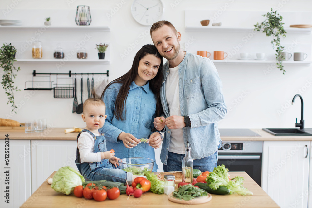 Portrait of joyful caucasian family of three posing in modern kitchen during cooking meals for breakfast. Curious daughter resting near salad bowl while smiling parents snuggling in dining room. Portrait of joyful caucasian family of three posing in modern kitchen during cooking meals for breakfast. Curious daughter resting near salad bowl while smiling parents snuggling in dining room.