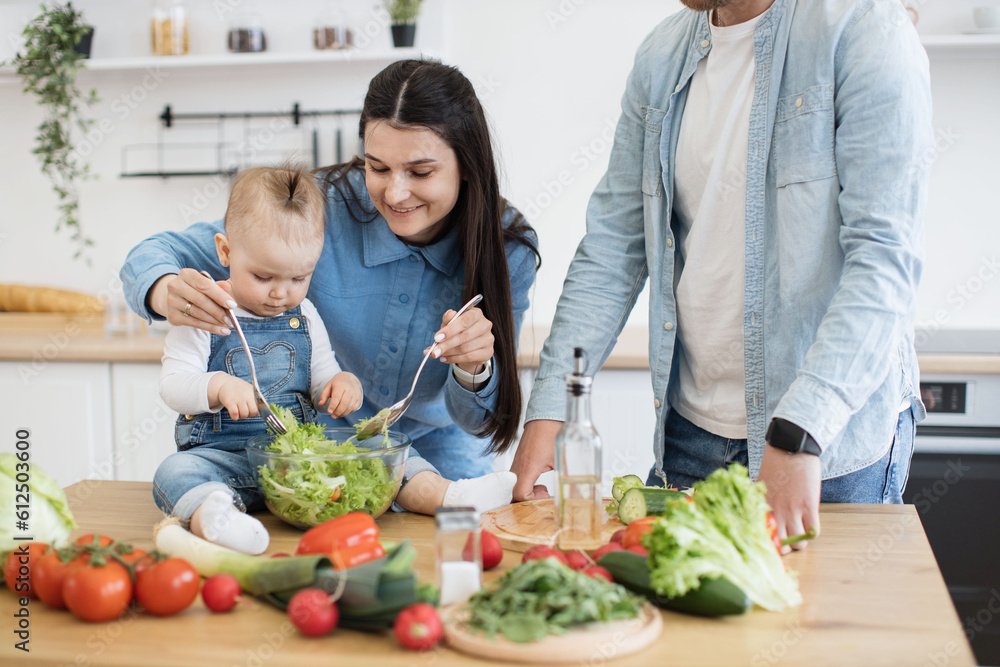 Cropped view of male in denim clothes leaning on wooden table covered with products and kitchenware. Attractive mother using spoons for salad mixture while active kid checking greens in bowl. Cropped view of male in denim clothes leaning on wooden table covered with products and kitchenware. Attractive mother using spoons for salad mixture while active kid checking greens in bowl.