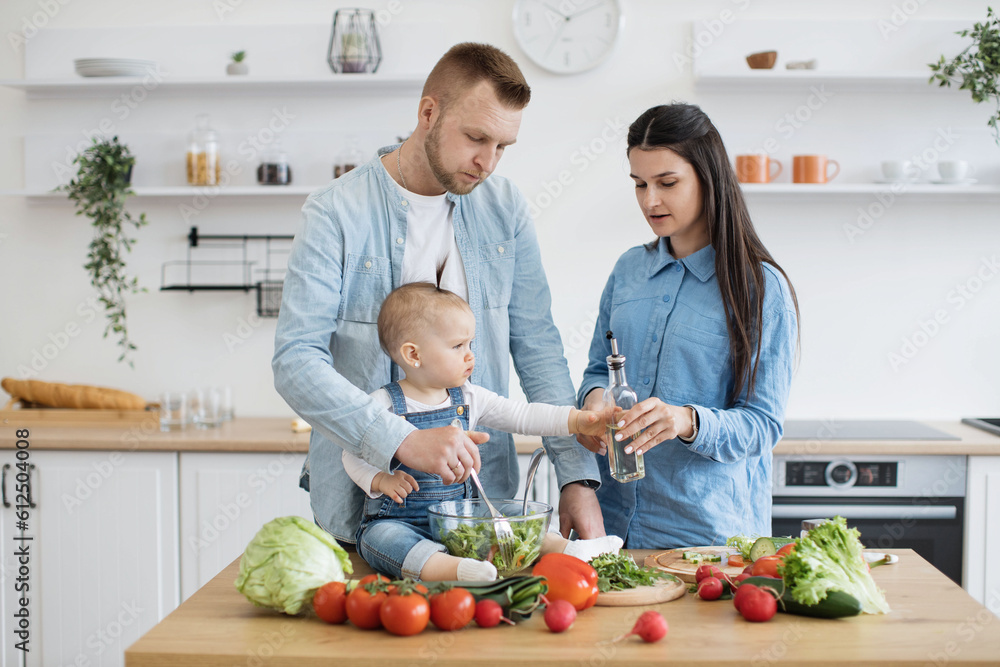 Mindful young woman adding olive oil to ingredients in bowl while her husband assisting with mixing components. Affectionate three-person family mastering culinary skills with tasty appetizer. Mindful young woman adding olive oil to ingredients in bowl while her husband assisting with mixing components. Affectionate three-person family mastering culinary skills with tasty appetizer.