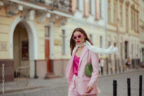 Fashionable elegant confident woman wearing trendy pink sunglasses, suit blazer, white silk scarf, trousers, with green shoulder bag, walking in street of European city. Copy, empty space for text