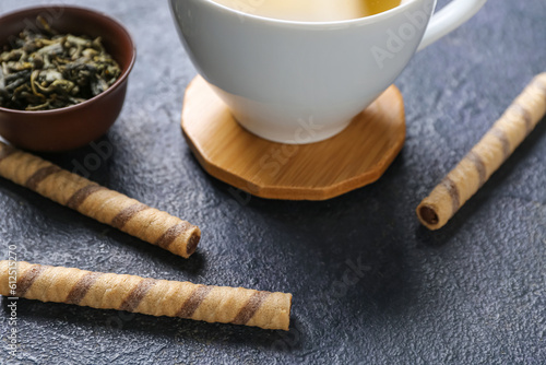 Drink coasters with cup of tea, dry leaves in bowl and wafer rolls on dark table