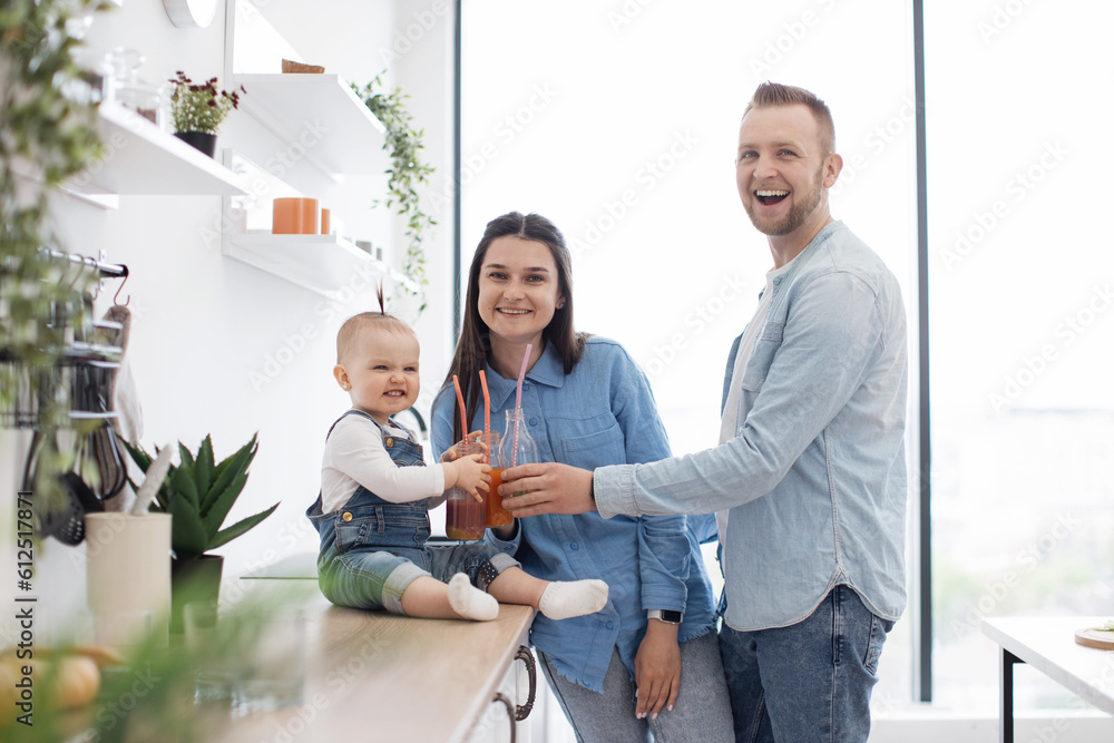 Fototapeta premium Portrait of two adults and child toasting glasses with fruit juice and smiling at camera. Cute caucasian family enjoying homemade fresh smoothies during healthy diet at home.