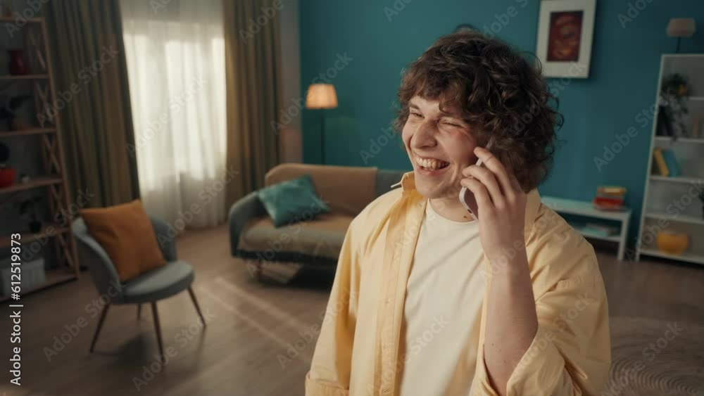 A young man speaks on a smartphone, tells the interlocutor about his housing, pointing with his index finger at the furniture. Portrait of a curlyhaired man in the living room close up. A young man speaks on a smartphone, tells the interlocutor about his housing, pointing with his index finger at the furniture. Portrait of a curlyhaired man in the living room close up.