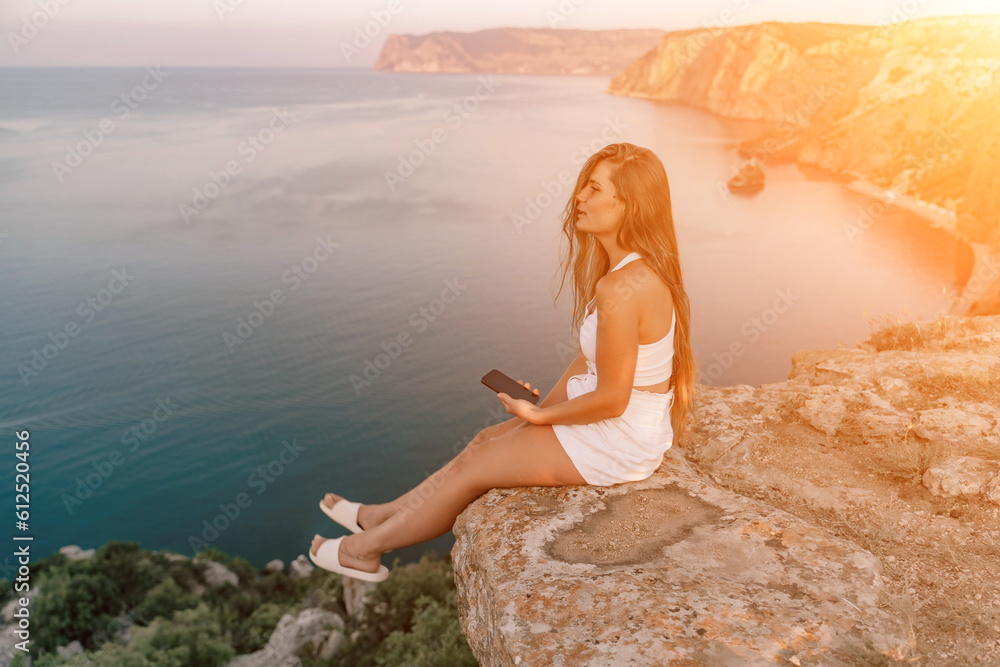 Happy woman in white shorts and T-shirt, with long hair, talking on the phone while enjoying the scenic view of the sea in the background.