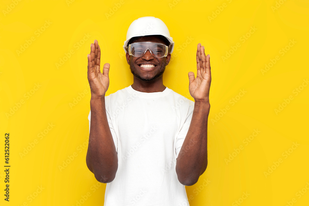 african american male engineer in hard hat and goggles holds his hands in front of him on yellow background