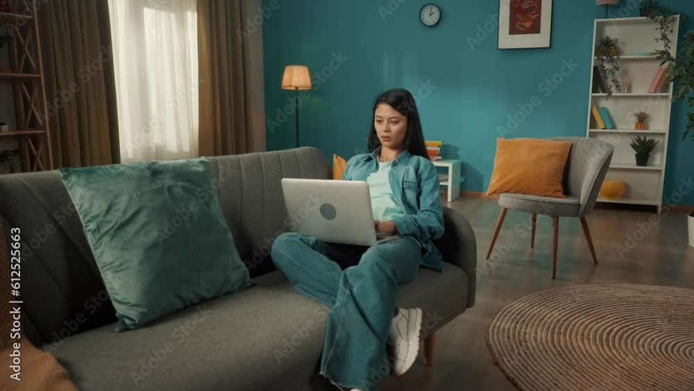 An attractive Asian woman sits on a sofa in the living room with a laptop on her lap. A woman is working, typing a text, a message, chatting. Remote work, freelancer.