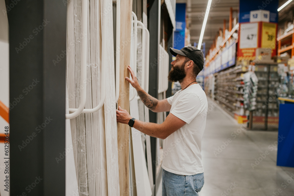 A young handsome man with a beard in casual clothes in a construction hypermarket in the lumber department selects wood building materials for the renovation of his house. - obrazy, fototapety, plakaty 