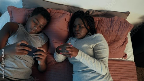 brazilian black child playing on smartphone lying in bed with his mother on mother's day and her tickling him photo