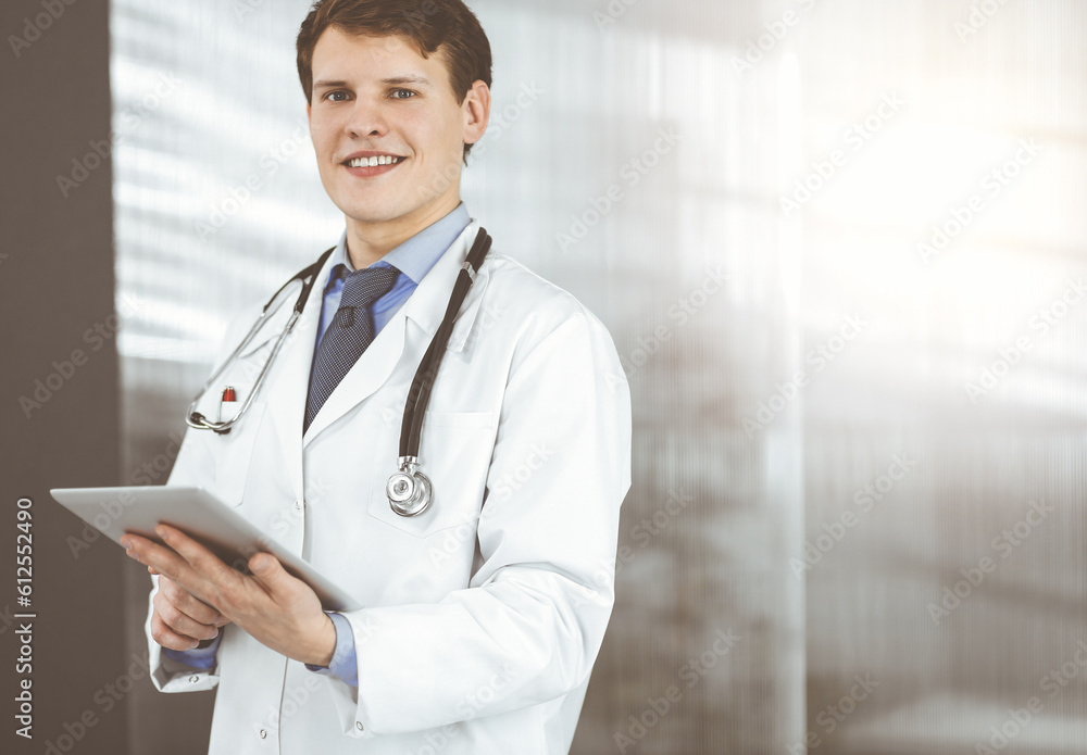 Young friendly doctor is checking some information on his computer tablet. Portrait of a professional physician at work in sunny clinic. Young friendly doctor is checking some information on his computer tablet. Portrait of a professional physician at work in sunny clinic.