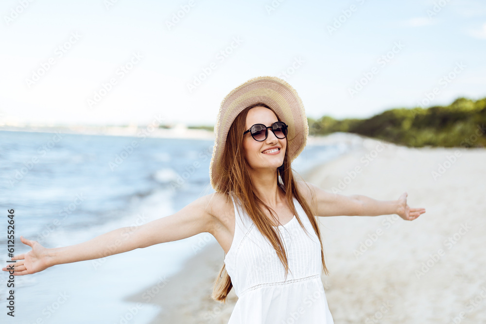Happy smiling woman in free happiness bliss on ocean beach standing with a hat, sunglasses, and open hands. Portrait of a multicultural female model in white summer dress enjoying nature during travel Happy smiling woman in free happiness bliss on ocean beach standing with a hat, sunglasses, and open hands. Portrait of a multicultural female model in white summer dress enjoying nature during travel