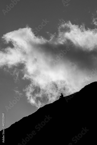 Vertical grayscale of a person sitting on the slope of a mountain against a dramatic sky