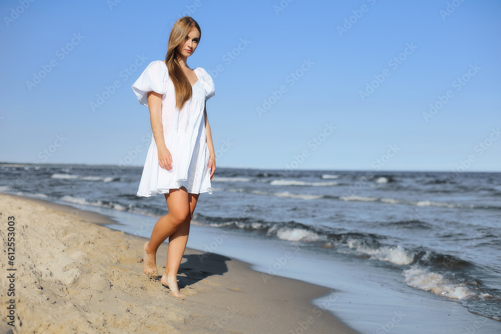 Happy smiling beautiful woman is walking on the ocean beach in a white summer dress.