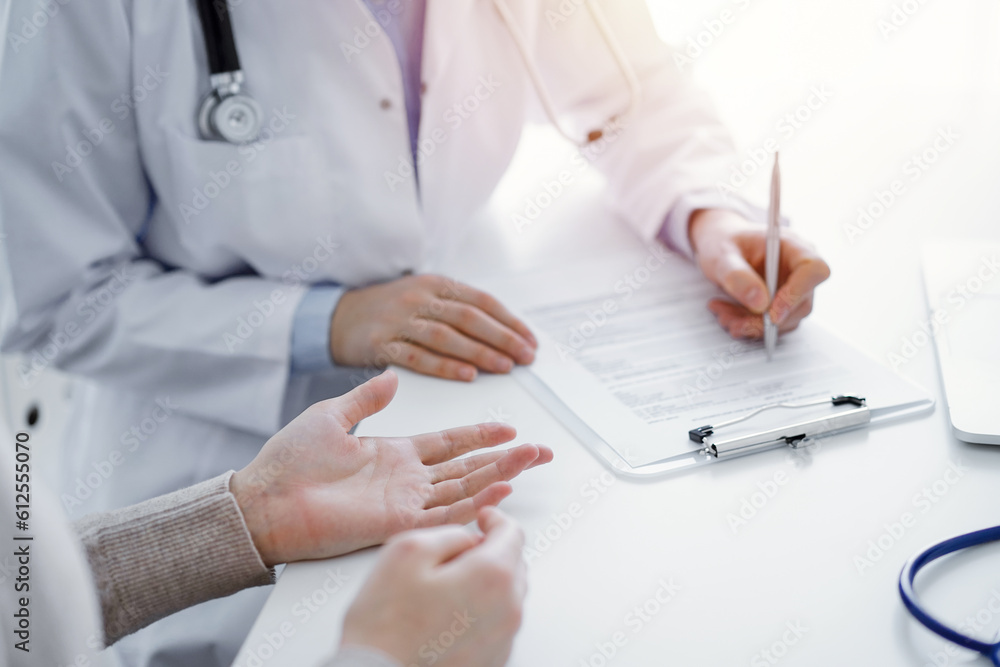Doctor and patient sitting at the table in clinic. The focus is on female physician's hands filling up the medication history record form or checklist, close up. Medicine concept. Doctor and patient sitting at the table in clinic. The focus is on female physician's hands filling up the medication history record form or checklist, close up. Medicine concept.