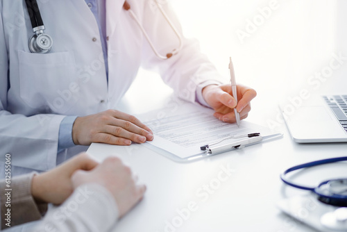 Doctor and patient sitting at the table in clinic. The focus is on female physician's hands filling up the medication history record form or checklist, close up. Medicine concept.