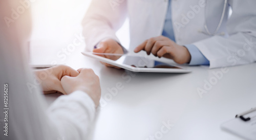 Doctor and patient sitting opposite each other at the desk in clinic. The focus is on female physician's hands using tablet computer, close up. Medicine concept.