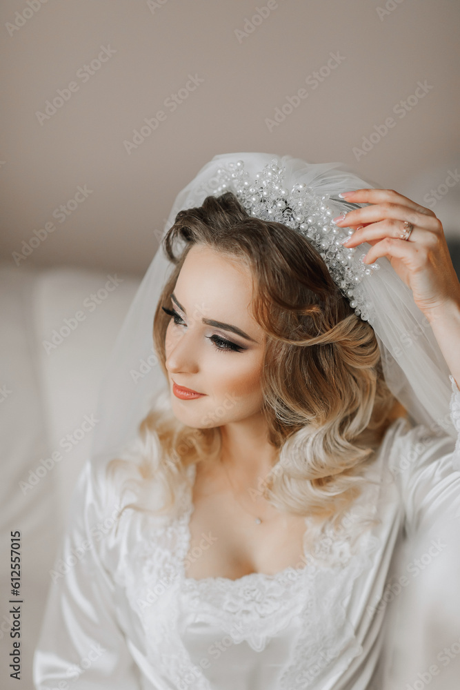 A blonde bride is wearing a satin robe and sitting in her room, posing and showing off her tiara and veil. Beautiful hair and make-up, open bust. Wedding portrait. A blonde bride is wearing a satin robe and sitting in her room, posing and showing off her tiara and veil. Beautiful hair and make-up, open bust. Wedding portrait.