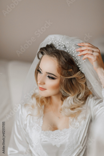 A blonde bride is wearing a satin robe and sitting in her room, posing and showing off her tiara and veil. Beautiful hair and make-up, open bust. Wedding portrait.