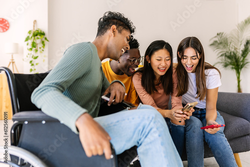Young group of people using mobile phone device sitting together on sofa at home. Inclusion and diversity concept with young indian man in wheelchair having fun with friends watching content on phone.