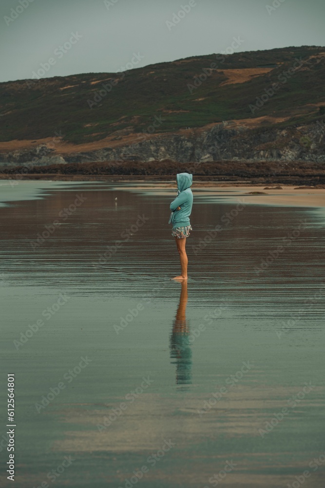 Naklejka premium Vertical shot of a female in a blue hoodie and shorts standing in the Woolacombe beach in July 2022