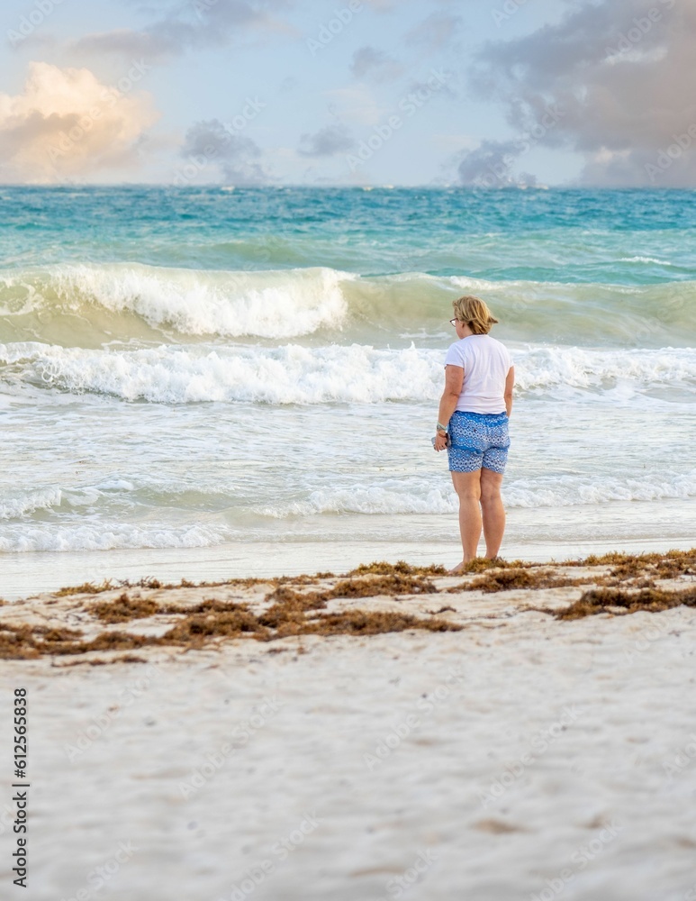 Vertical shot of a caucasian female standing on the beach shore looking at the wave Vertical shot of a caucasian female standing on the beach shore looking at the wave