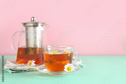 Teapot with cup of natural chamomile tea and flowers on turquoise table near pink wall