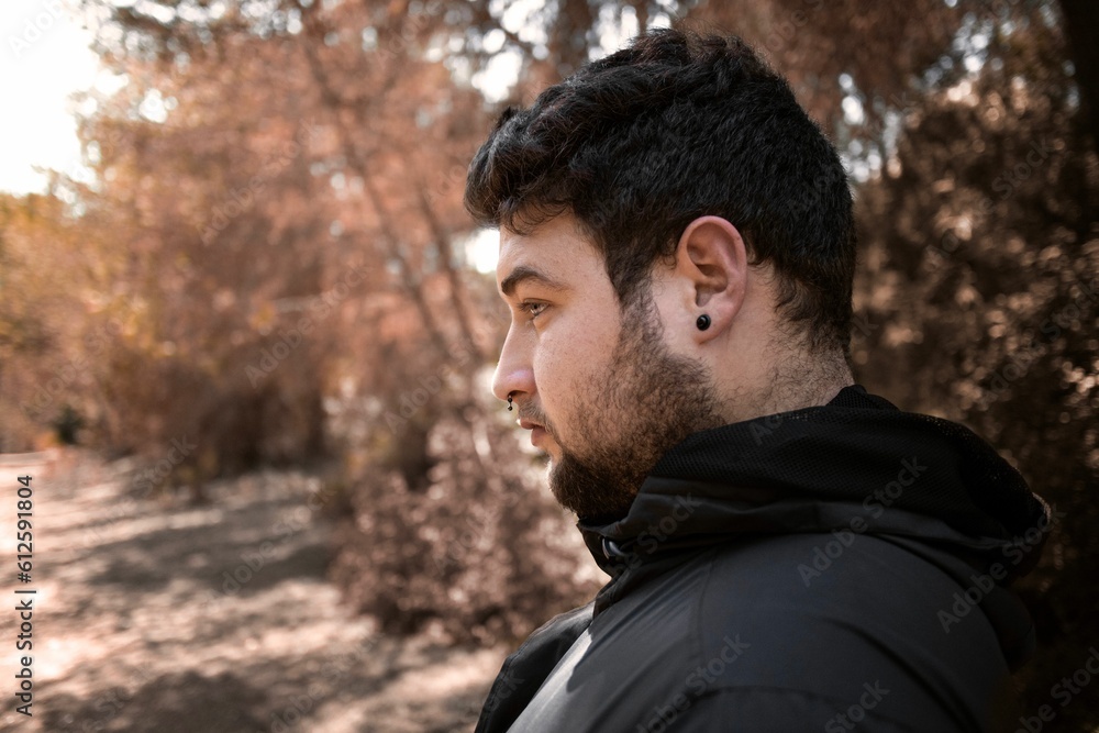 Side profile of a Caucasian man with a septum piercing with the fall trees in the blurred background