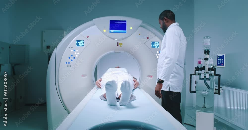 Patient after tomography procedure. Multicultural doctor finishes magnetic resonance imaging. Patient is lying at CT scanner bed and moving out of MRI capsule. Well-equipped tomography room. Patient after tomography procedure. Multicultural doctor finishes magnetic resonance imaging. Patient is lying at CT scanner bed and moving out of MRI capsule. Well-equipped tomography room.