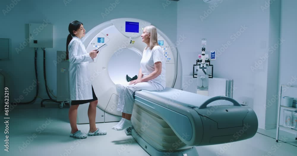 Female doctor gestures and points to MRI capsule. Female patient is sitting at CT scanner bed and listening to doctor attentively. Medical worker is holding tablet and talk to patient. Female doctor gestures and points to MRI capsule. Female patient is sitting at CT scanner bed and listening to doctor attentively. Medical worker is holding tablet and talk to patient.