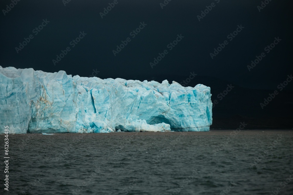 Large iceberg with broken off pieces seen against a backdrop of dark, cloudy sky Large iceberg with broken off pieces seen against a backdrop of dark, cloudy sky