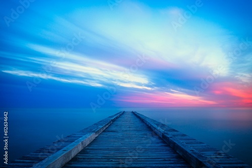 Fototapeta Long wooden pier extends out into a tranquil body of water during the pink and blue sunset