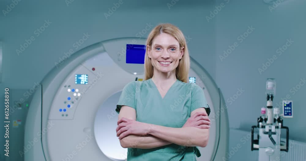 Portrait of female doctor at background of MRI capsule. Middle-aged doctor in blue gown is smiling with teeth and posing for camera. Woman doctor with arms crossed is looking straight at camera. Portrait of female doctor at background of MRI capsule. Middle-aged doctor in blue gown is smiling with teeth and posing for camera. Woman doctor with arms crossed is looking straight at camera.