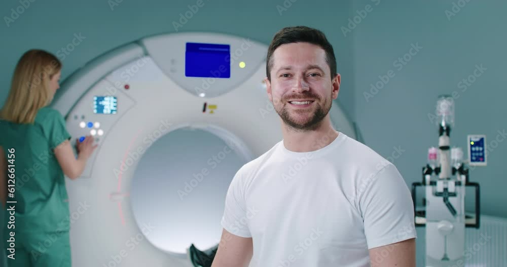 Happy male patient at well-equipped MRI laboratory, Portrait of cheerful man at modern tomography room. Man smiles with teeth at background of MRI capsule. Female doctor adjusts CT scanner behind man. Happy male patient at well-equipped MRI laboratory, Portrait of cheerful man at modern tomography room. Man smiles with teeth at background of MRI capsule. Female doctor adjusts CT scanner behind man.