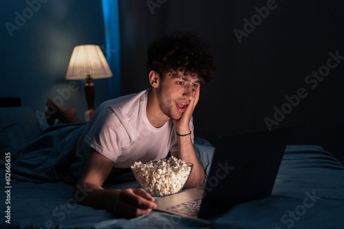 Shocked young male eating popcorn and watching film on laptop while resting on bed at night at home