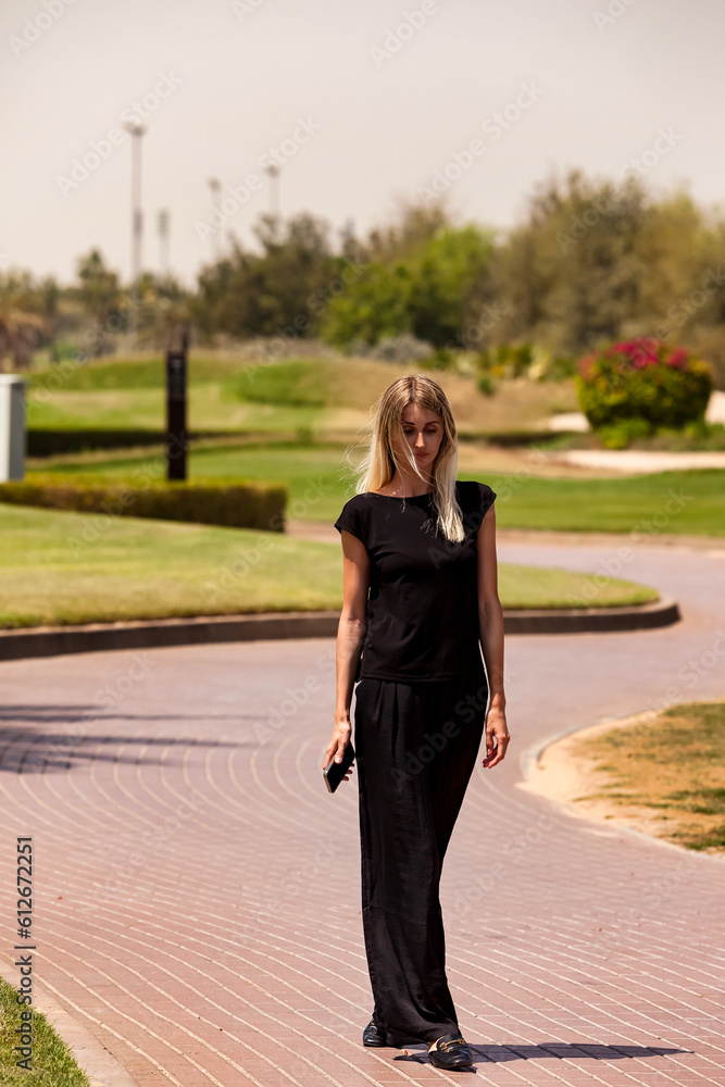 Full length of stylish thoughtful pretty woman in black clothes walking in urban park outdoors at greenery. Pensive lovely young lady posing in nature. Business activity concept. Copy ad text space