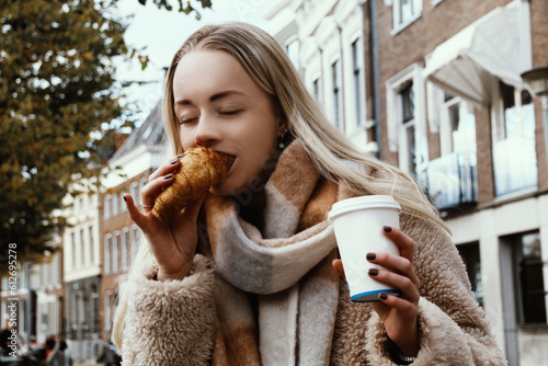 Young blond woman with croissant drinking coffee on a street