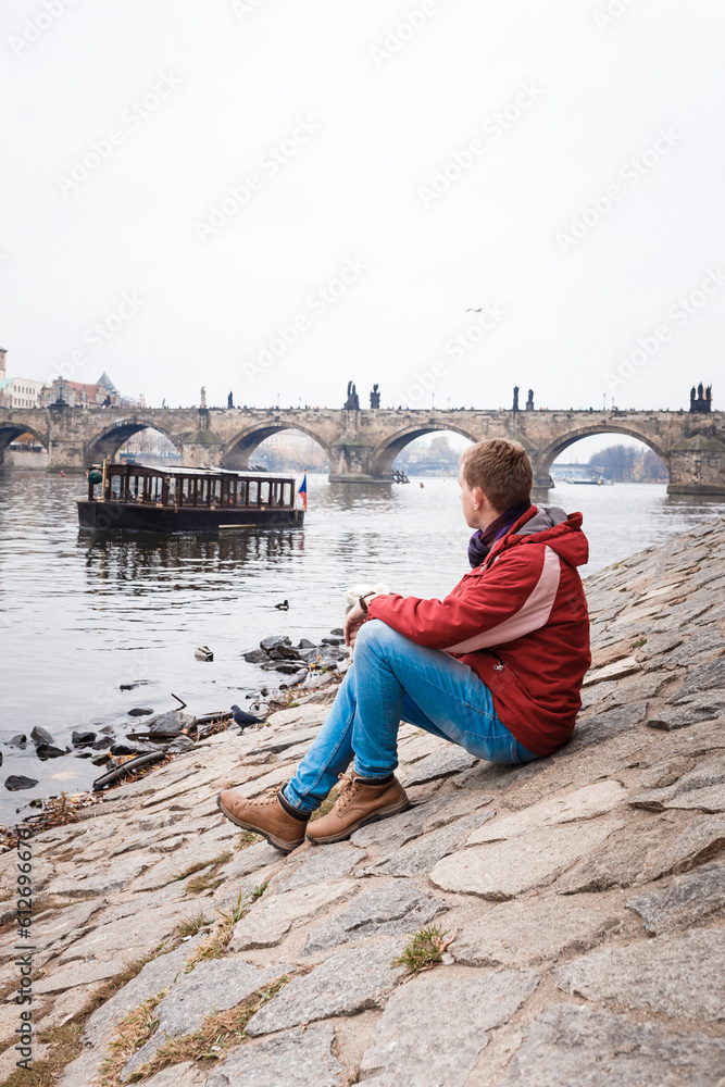 male tourist in red jacket, blue jeans on embankment of Prague