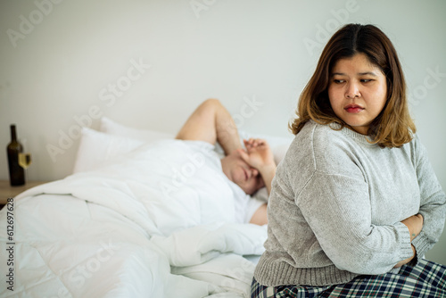 Young unhappy married couple in bedroom. Woman sitting on bed feel boring in relationship with her husband sleep at bedroom. Party every day and drink alcohol heavily.