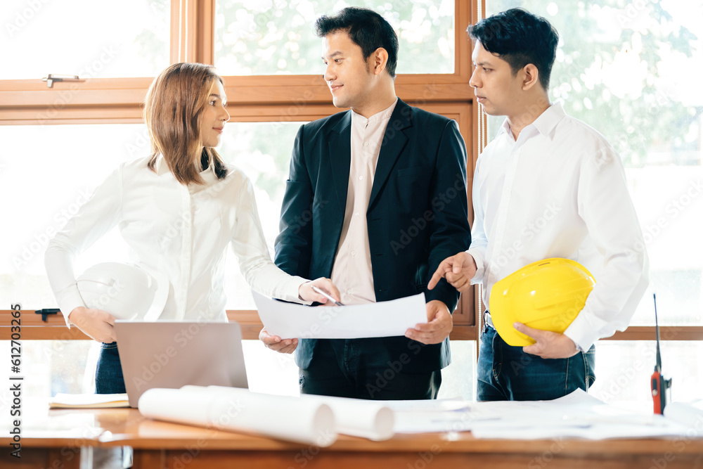 Three Asian male and female architects are meeting for a structural engineering design project. Blueprints in the office, a desk, a computer, a walkie-talkie, a helmet, and wearing a uniform. Three Asian male and female architects are meeting for a structural engineering design project. Blueprints in the office, a desk, a computer, a walkie-talkie, a helmet, and wearing a uniform.
