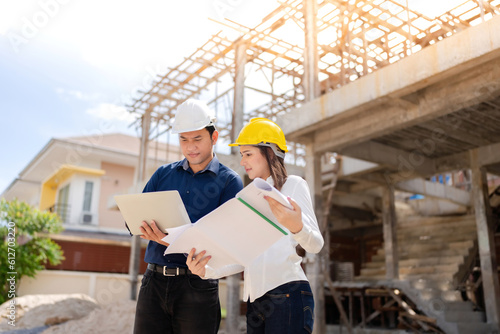 A team of 2 Asian male and female engineers, mechanic managers and staff. Business meeting with tablet and blueprint In construction site, architecture industry, holding listnote, wearing vest, helmet