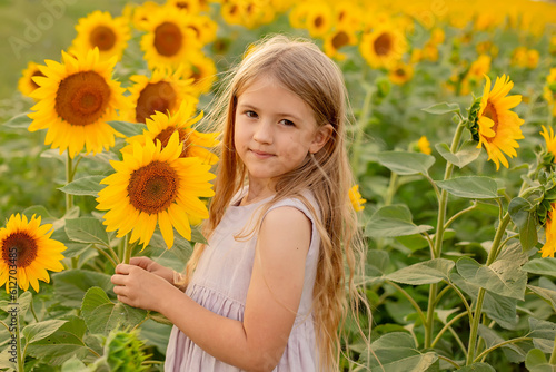 a blonde girl with long hair in a pink linen dress stands in a field with sunflowers and holds a flower in her hands