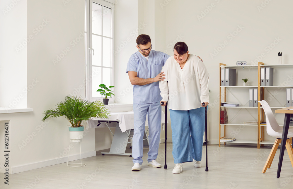 A young cheerful friendly nurse helping fat young patient to walk with her crutches in medical clinic. Male physiotherapist or health care worker helping overweight woman in rehab. A young cheerful friendly nurse helping fat young patient to walk with her crutches in medical clinic. Male physiotherapist or health care worker helping overweight woman in rehab.