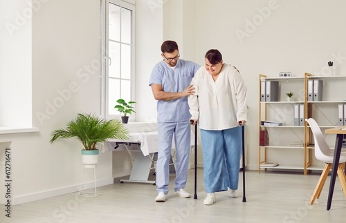 Fotografia A young cheerful friendly nurse helping fat young patient to walk with her crutches in medical clinic