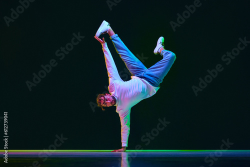 Dynamic image of talented, active young man in sport style clothes dancing breakdance against black studio background in neon light