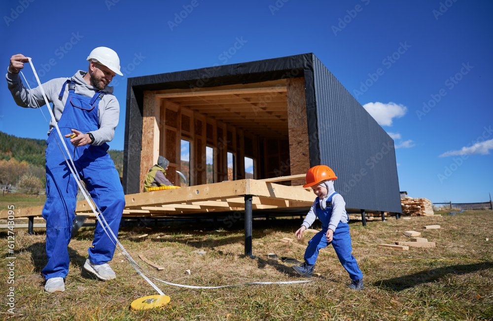 Father with toddler son building wooden frame house in the Scandinavian style barnhouse. Male builder and kid playing with tape measure on construction site on sunny day. Carpentry and family concept. Father with toddler son building wooden frame house in the Scandinavian style barnhouse. Male builder and kid playing with tape measure on construction site on sunny day. Carpentry and family concept.