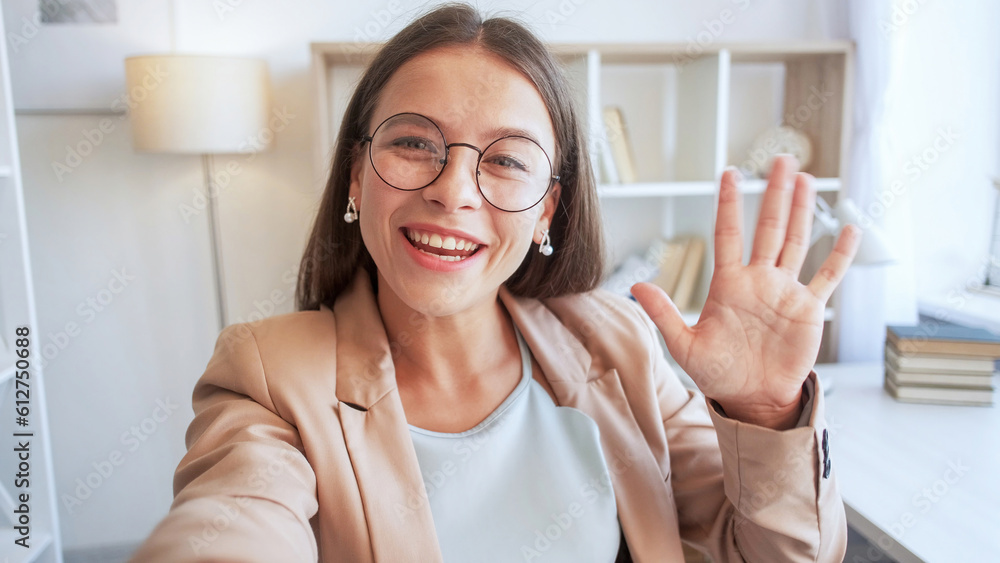 Video blog. Influencer streaming. Beautiful happy expressive young woman with big smile in joyful mood in glasses waving hand to camera for warm greeting in white room.
