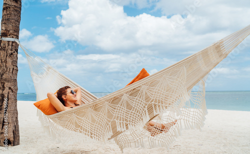 Young woman relaxing in wicker hammock on the sandy beach on Mauritius coast and enjoying wide ocean view waves. Exotic countries vacation and mental health concept image.