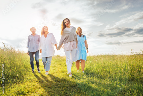 Portrait of four cheerful smiling women holding hand in hand walking by a high green grass meadow. They looking at the camera. Woman's friendship, relations, and happiness concept image.
