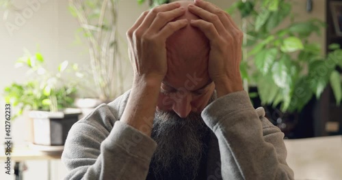 Worried 50 years old man with bald head and long beard holding hands on his head having deep thoughts at his home